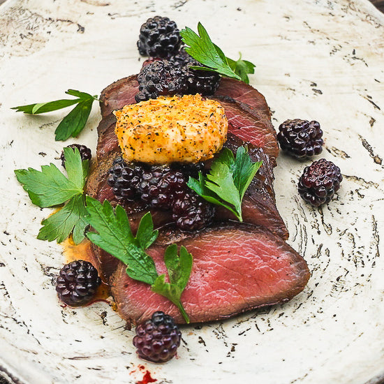 Sliced venison strip loin on a white plate topped with spiced butter and blackberries