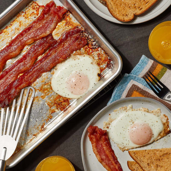 Sheet pan of bacon and eggs with one serving removed and toast and orange juice on the side