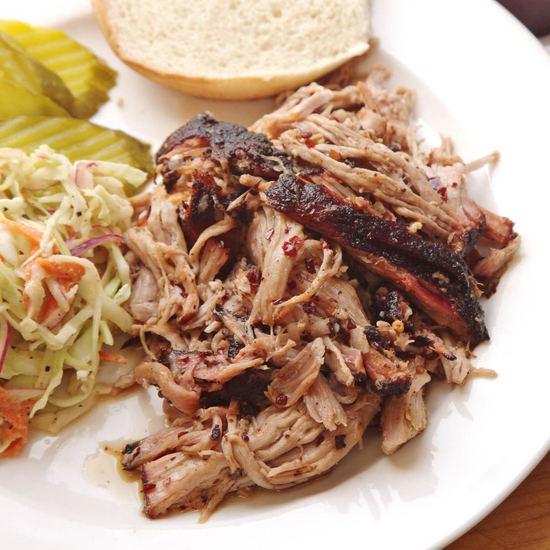 Shredded barbecue pork shoulder on a white plate with coleslaw, pickles, and a bun on the side